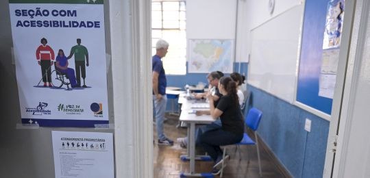 Foto mostra cartaz com a identificação seção com acessibilidade ao lado da porta de uma sala