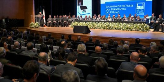 Foto mostra um teatro lotado. Ao fundo, diversas pessoas sentadas junto a uma mesa sobre o palco.