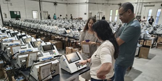 Foto mostra dezenas de urnas eletrônicas sobre bancadas, dentro de um galpão. Algumas pessoas es...