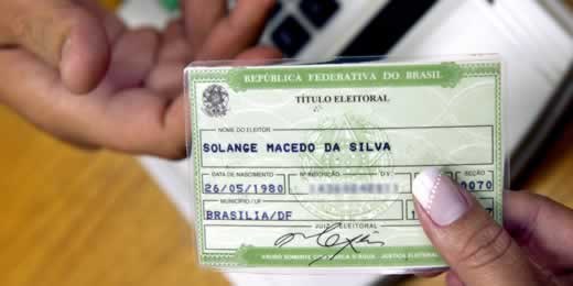 Mão feminina passando título eleitoral para mão masculina, com leitor biométrico ao fundo.