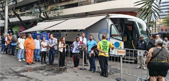 Foto mostra uma fila de pessoas junto a um ônibus com a identidade visual TRE Aqui
