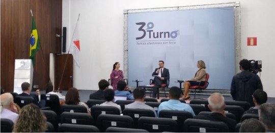 Debatedoras e mediador estão sentados em cadeiras no palco do auditório do TRE-MG, na edição do ...