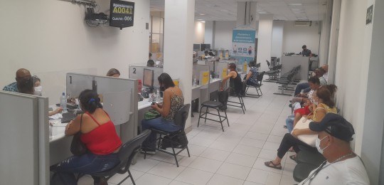 Pessoas sentadas aguardando atendimento e pessoas sentadas junto a guichês enquanto são atendidas.