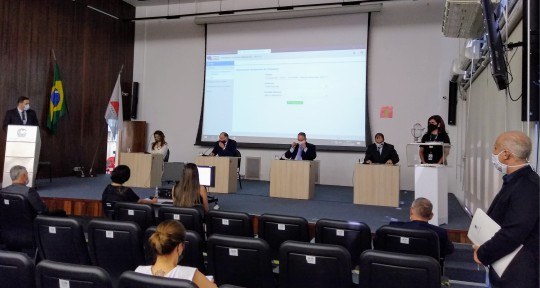 Foto mostra parte do auditório, com poucas pessoas sentadas na plateia. Uma mulher e três homens...