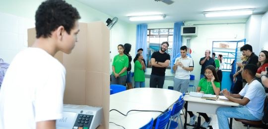 TRE-MG-camara-mirim-2024-1 Foto de pessoas em volta de uma mesa com um eleitor que votou em uma urna eletrônica
