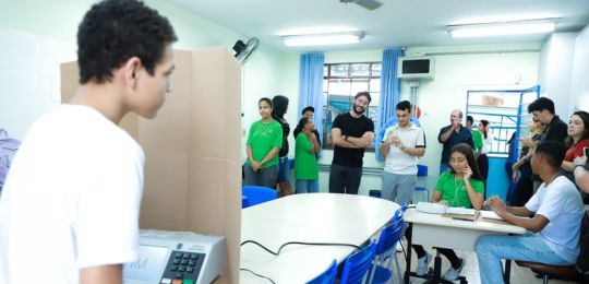 Foto de pessoas em volta de uma mesa com um eleitor que votou em uma urna eletrônica