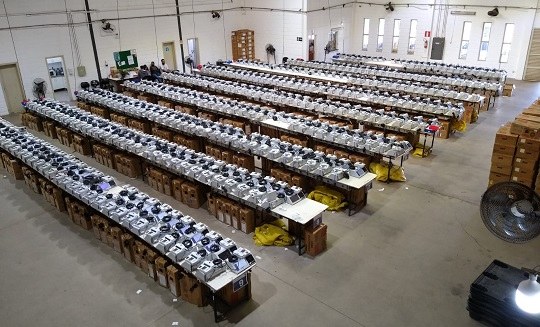 Galpão com dezenas de urnas eletrônicas sobre nove bancadas.