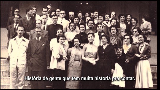 Foto em preto e branco que mostra várias pessoas de pé, posando para a câmera. 