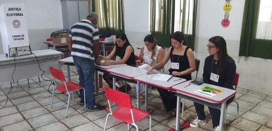 TRE-MG-eleicao-suplementar-martins-soares-2025 Imagem de uma seção eleitoral, contendo cinco mesas com quatro pessoas sentadas, atendendo um el...