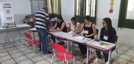 Imagem de uma seção eleitoral, contendo cinco mesas com quatro pessoas sentadas, atendendo um el...