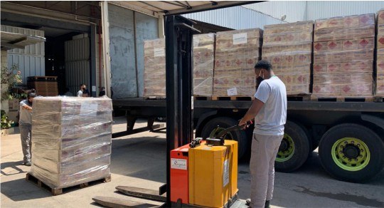 Foto mostra um homem empurrando um pallet com várias caixas envoltas em plástico. Outro homem ma...