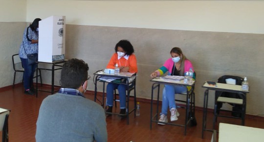 Foto de uma seção eleitoral. Uma eleitora está atrás da cabine de votação. Duas mesárias estão s...