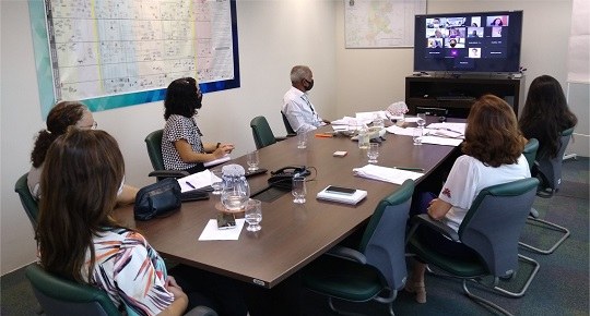 Foto mostra seis pessoas sentadas ao redor de uma mesa oval. Elas olham para uma televisão, em c...