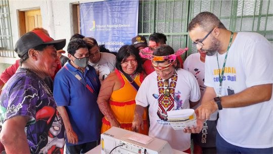 A imagem mostra um servidor do TRE auxiliando indígenas maxakali na operação de uma urna eletrôn...