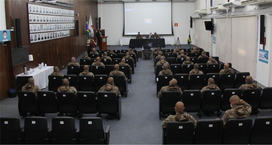 TRE-MG-palestra-pm-novembro Foto do auditório do TRE, mostrando policiais militares sentados na plateia e três homens sentad...