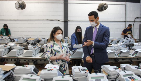 Uma mulher de vestido estampado e um homem de terno azul estão de pé perto de uma bancada com ur...