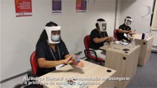 Foto que mostra três mesários sentados atrás de mesas de trabalho. Eles usam máscaras e protetor...
