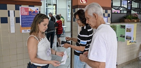 Um casal de idosos e uma mesária jovem na entrada de uma seção eleitoral. Dentro da sala, eleito...