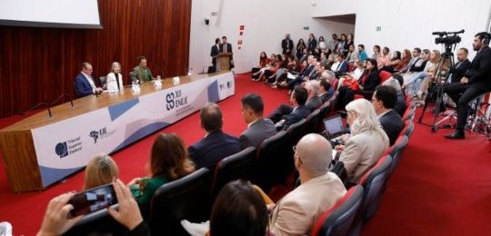 TRE-XII-Encontro-Nacional-das-Escolas-Judiciárias-Eleitorais Foto de um auditório com uma mesa a frente, onde se lê XII ENEJE, com pessoas sentadas à mesa e ...