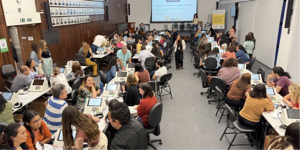 Foto mostra dezenas de pessoas dentro de um auditório, sentadas junto a mesas sobre as quais há ...