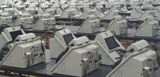 urnas eletrônicas TRE-MG Foto mostra dezenas de urnas eletrônicas modelo 2022 sobre bancadas de madeira.