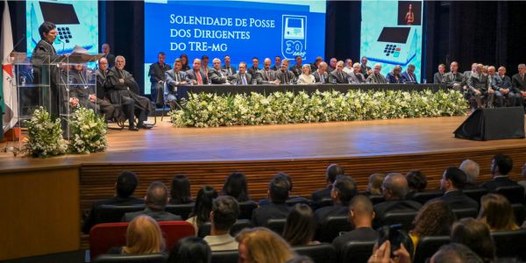 Foto mostra um teatro lotado. Ao fundo, diversas pessoas sentadas junto a uma mesa sobre o palco.