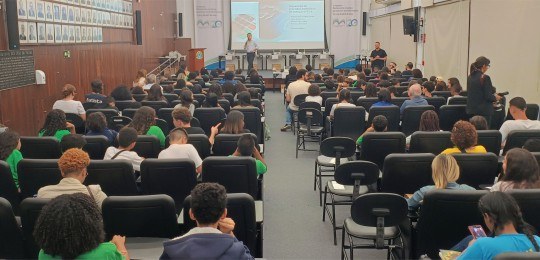 Foto mostra um auditório lotado de estudantes. Ao fundo, o servidor do TRE Glaysson Rocha está d...