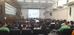 Foto de um auditório com dezenas de adolescentes na plateia. Ao fundo, no palco, uma mulher negr...