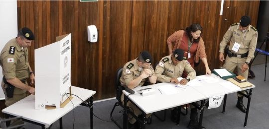 Um policial militar vota na urna eletrônica, enquanto outros três atuam como mesários.