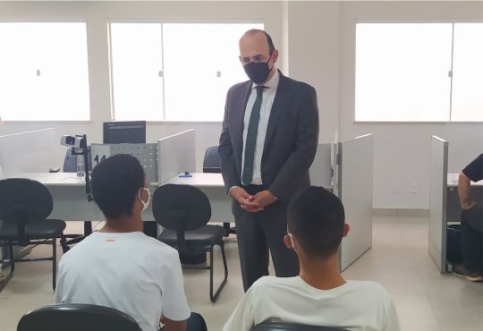 A imagem mostra um homem de terno em pé conversando com dois jovens sentados, em uma Central de ...