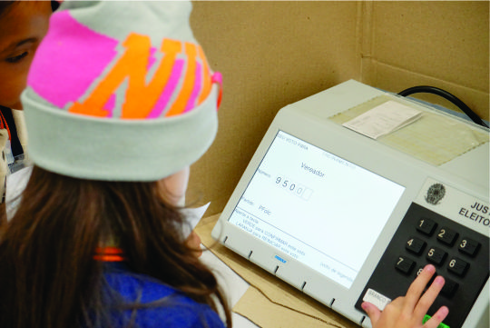 A imagem mostra uma criança de costas votando em uma urna eletrônica.